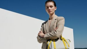 ミニマルな白い壁と澄み切った深い青空を背景にポーズをとるファッショナブルな女性。女性は白いトップスの上にスタイリッシュなベージュのブレザーを羽織り、自信たっぷりに腕を組んでいる。黄色のストラップが印象的な、なめらかな淡い色のハンドバッグを持っている。